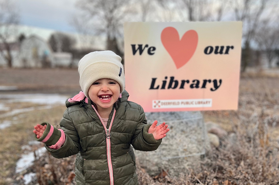 A little girl poses in front of a Deerfield Public Library yard sign.